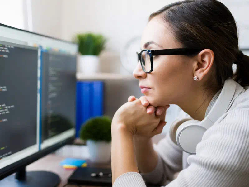 Woman wearing glasses and headphones studying code on dual monitors
